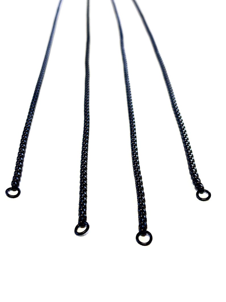 May include: Four black metal chain necklaces with circular clasps are displayed against a white background. The chains are made of small, interlocking links. The chains are evenly spaced and angled towards the viewer.