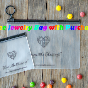 May include: Two clear plastic zippered bags with black zippers and black trim. The bags are on a wooden surface. The larger bag has the text "Shiny Little Blessings" printed on it. The smaller bag has the text "Free Jewelry Bag with Purchase" printed on it.