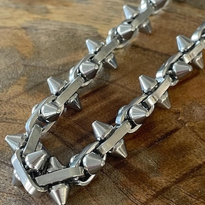 May include: A silver chain necklace featuring cone-shaped spikes. The chain links are rectangular and interlock. Spikes are evenly spaced along the chain. The necklace is displayed on a wooden surface.
