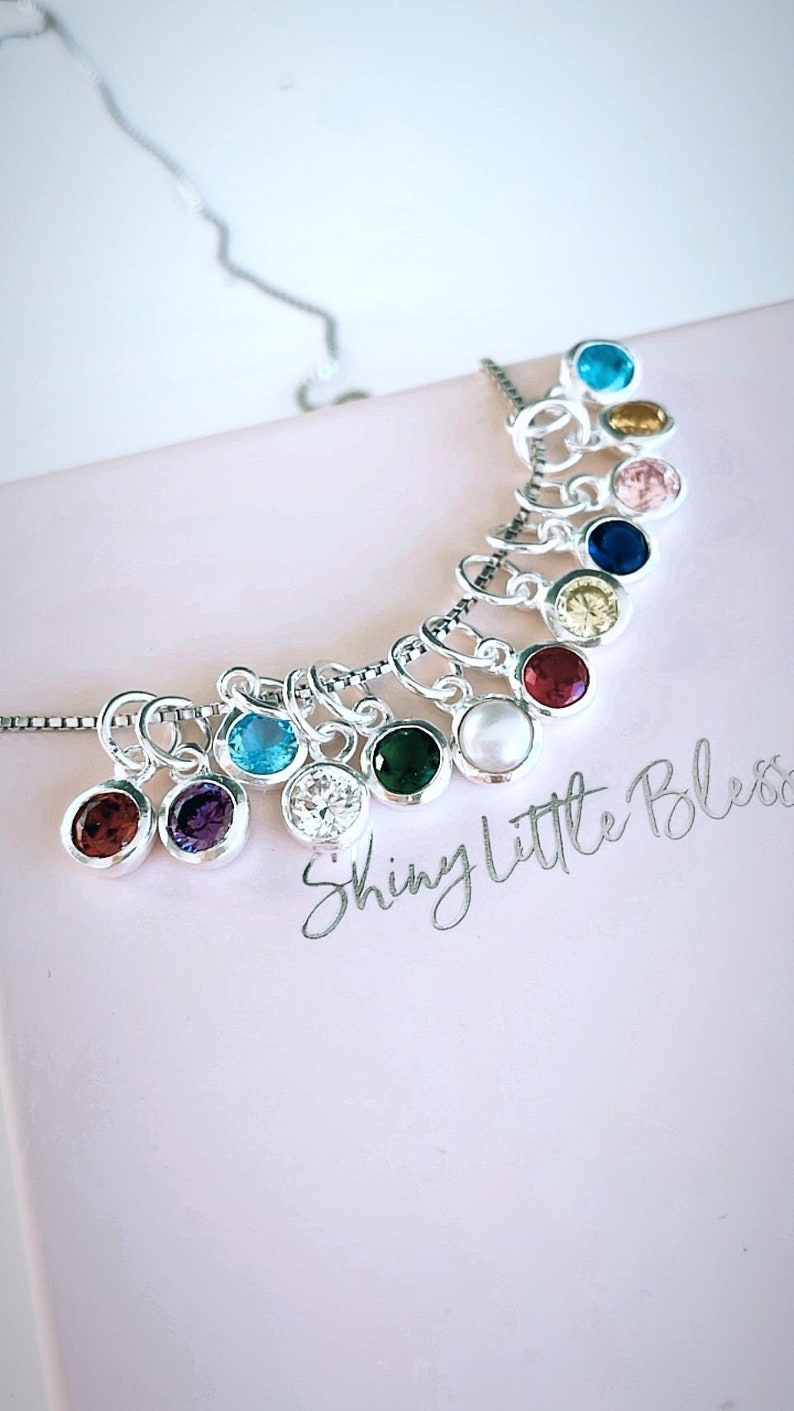 May include: A silver necklace with multiple charms, each featuring a different colored gemstone. The charms are arranged in a semi-circle shape, with the text "Shiny Little Bless" written on the surface below.