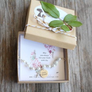 May include: A white pearl bracelet with a silver charm that says "Queen Kendall" in a gold font. The bracelet is in a small brown box with a white and gold ribbon. The box has a card inside with the text "May you always live your faith with joy."