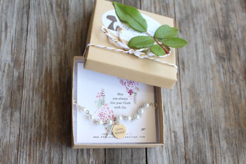 May include: A white pearl bracelet with a silver charm that says "Queen Kendall" in a gold font. The bracelet is in a small brown box with a white and gold ribbon. The box has a card inside with the text "May you always live your faith with joy."