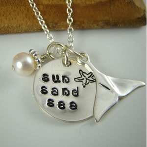 Puede incluir: Un collar de plata con un colgante redondo estampado con las palabras sun sand sea, un grabado de estrella de mar, un dije de cola de ballena de plata y una perla blanca individual, todo unido a una fina cadena de eslabones.
