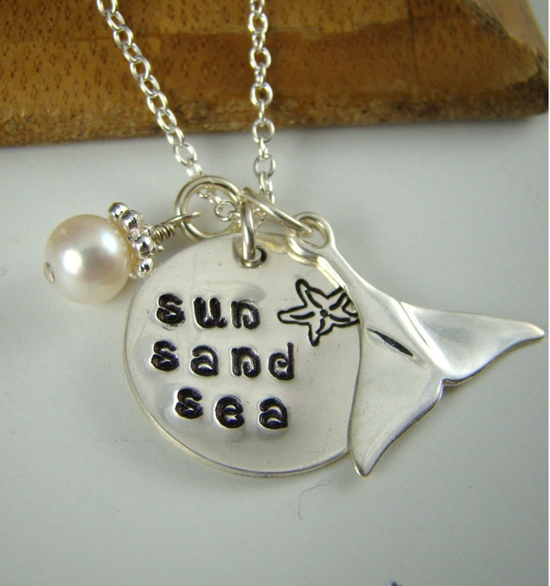 Puede incluir: Un collar de plata con un colgante redondo estampado con las palabras sun sand sea, un grabado de estrella de mar, un dije de cola de ballena de plata y una perla blanca individual, todo unido a una fina cadena de eslabones.
