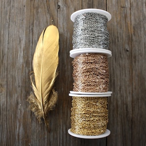 May include: Three spools of chain, one silver, one rose gold, and one gold, are stacked on top of each other on a wooden surface. A gold feather is to the left of the spools.
