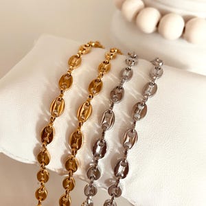 Op de afbeelding: Twee gouden en zilveren kettingen met een ankerketting ontwerp. De kettingen liggen op een wit oppervlak.