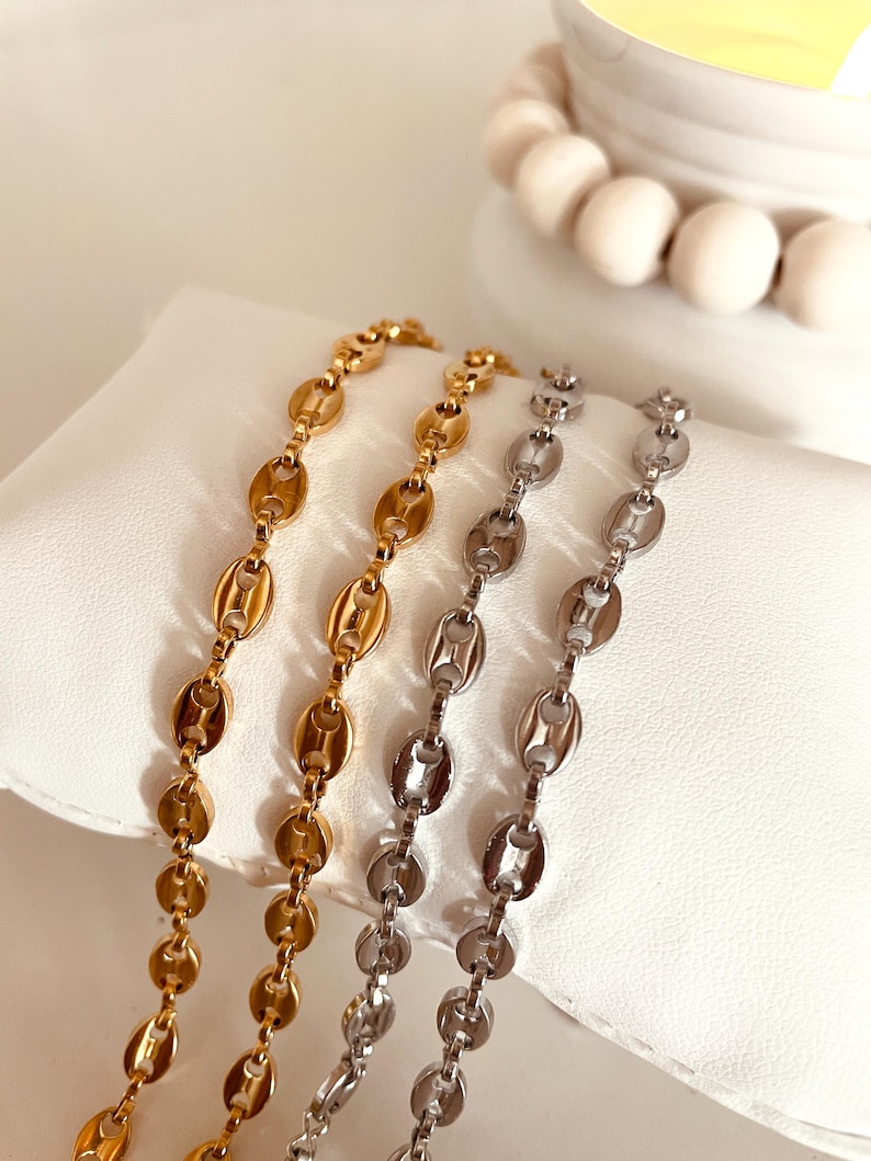 Op de afbeelding: Twee gouden en zilveren kettingen met een ankerketting ontwerp. De kettingen liggen op een wit oppervlak.