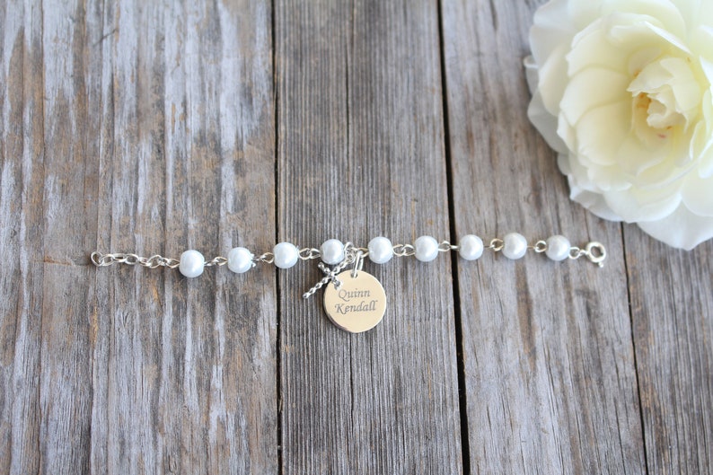 May include: A white pearl bracelet with a silver chain and a silver charm with the name "Quinn Kendall" engraved on it.