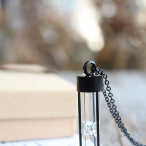 May include: A black hourglass necklace with a silver chain. The hourglass is made of glass and has a black metal frame. The necklace is on a white surface with the text "HANDCRAFTED WITH LOVE" visible.