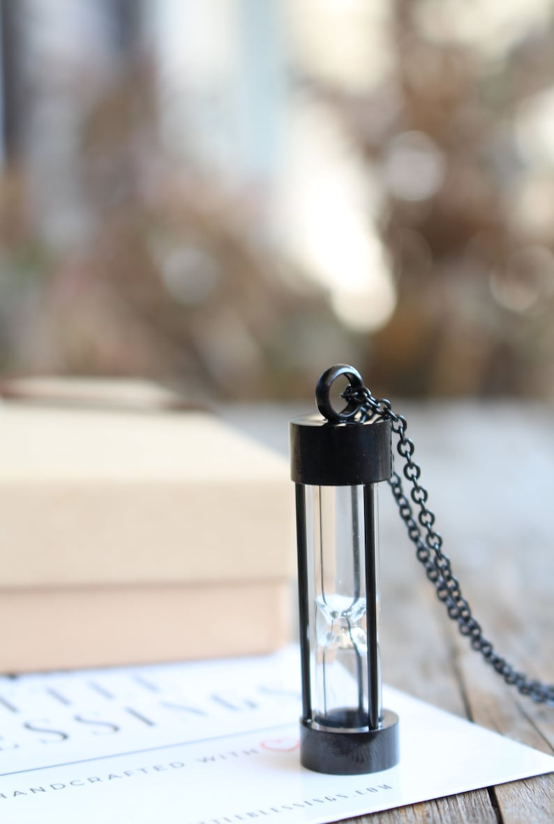 May include: A black hourglass necklace with a silver chain. The hourglass is made of glass and has a black metal frame. The necklace is on a white surface with the text "HANDCRAFTED WITH LOVE" visible.