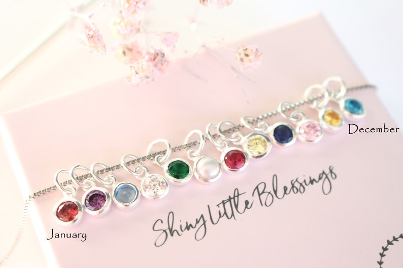 May include: A silver necklace with charms featuring different colored gemstones. The charms are shaped like hearts and have a small loop at the top. The necklace is displayed on a pink background with the text "Shiny Little Blessings" and the months "January" and "December".