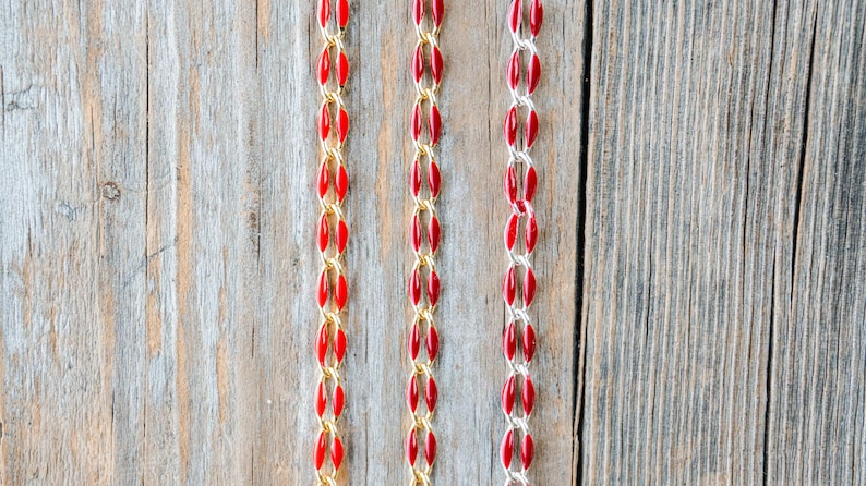 May include: Three strands of red and gold chain on a wooden background. The chains are made of small, oval-shaped beads.