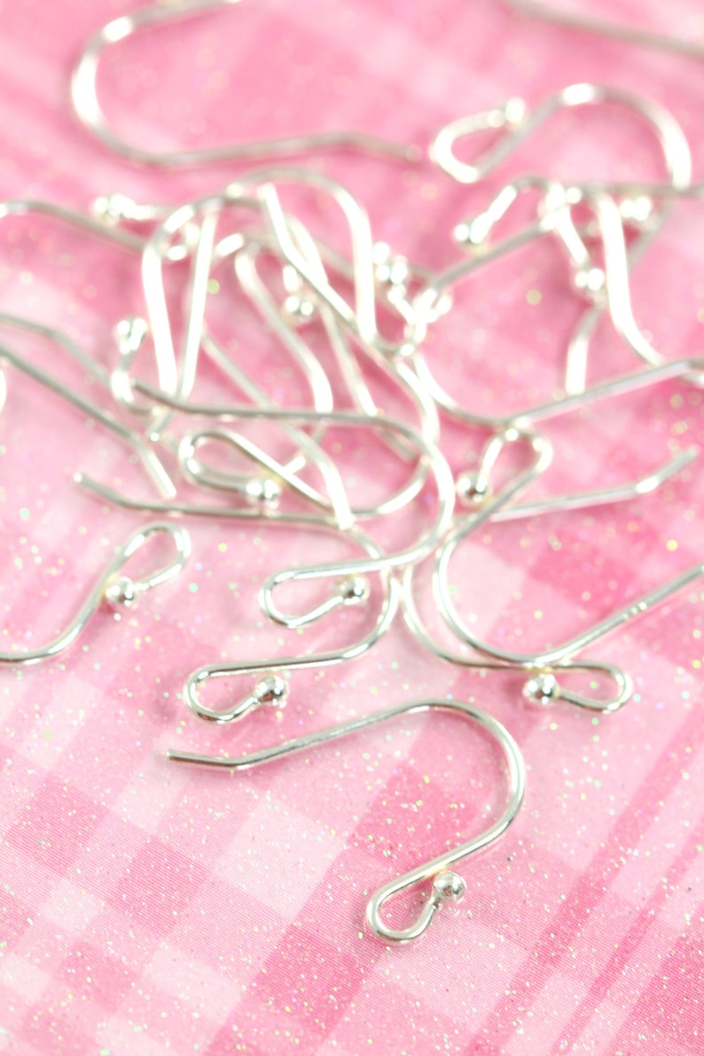 May include: A close-up of a pile of silver earring hooks on a pink sparkly background.