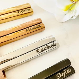 May include: Four pairs of chopsticks with personalized names: Cora, Malia, Rachel, and Skyla. The chopsticks are made of different metals, including gold, rose gold, silver, and black.