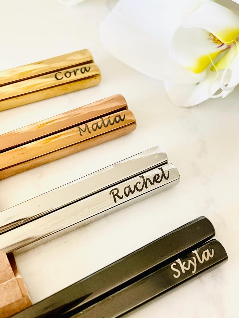 May include: Four pairs of chopsticks with personalized names: Cora, Malia, Rachel, and Skyla. The chopsticks are made of different metals, including gold, rose gold, silver, and black.