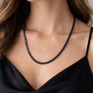 Op de afbeelding: Een zwarte ketting met vierkante schakels. De ketting bestaat uit kleine, vierkante schakels, waardoor een strak en modern ontwerp ontstaat. De ketting wordt gedragen over een zwarte satijnen top.