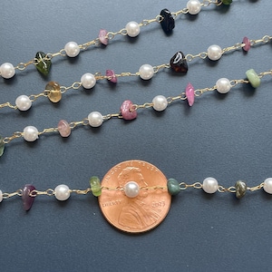 Puede incluir: Primer plano de varias hebras de delicados collares. Cada collar presenta una cadena dorada con perlas blancas y piedras preciosas de colores de forma irregular. Se incluye una moneda como referencia de tama&ntilde;o.