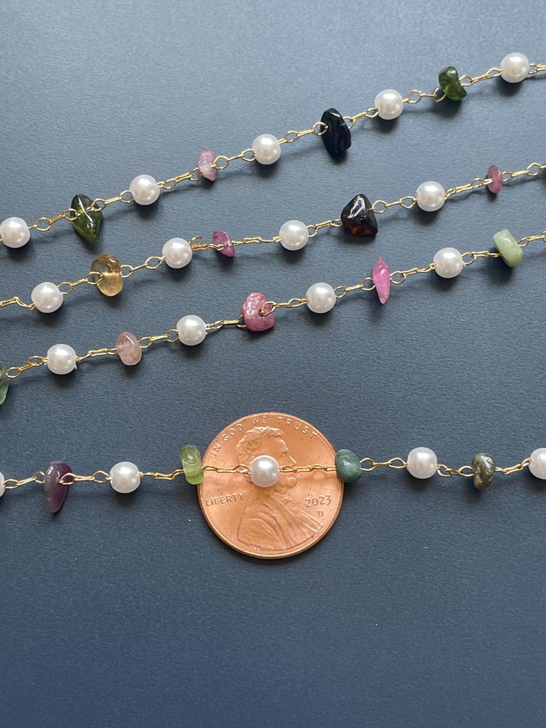 Puede incluir: Primer plano de varias hebras de delicados collares. Cada collar presenta una cadena dorada con perlas blancas y piedras preciosas de colores de forma irregular. Se incluye una moneda como referencia de tama&ntilde;o.