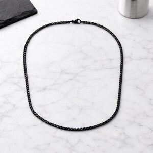 May include: A black chain necklace with a box link design. The necklace is made of a dark metal and is displayed on a white and grey marble surface. The necklace is a simple, yet elegant piece of jewellery.