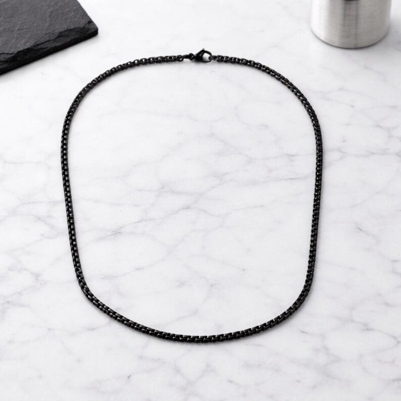 May include: A black chain necklace with a box link design. The necklace is made of a dark metal and is displayed on a white and grey marble surface. The necklace is a simple, yet elegant piece of jewellery.