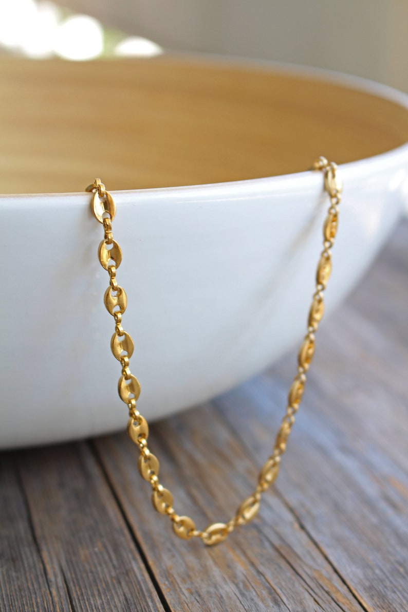 May include: A gold chain necklace with a mariner link design. The chain is laying on a wooden surface next to a white bowl.