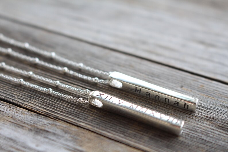 May include: Two silver bar necklaces with delicate chains. One bar has the name "Hannah" engraved on it, and the other has Roman numerals "XII V MMXV" engraved on it.