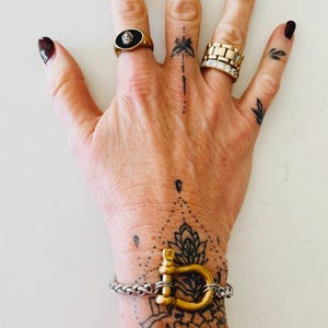 Pu&ograve; includere: Un braccialetto d'oro con una grande chiusura a forma di U in oro. Il braccialetto &egrave; fatto di una catena d'argento. Il braccialetto &egrave; al polso di una persona, che ha diversi piccoli tatuaggi neri.