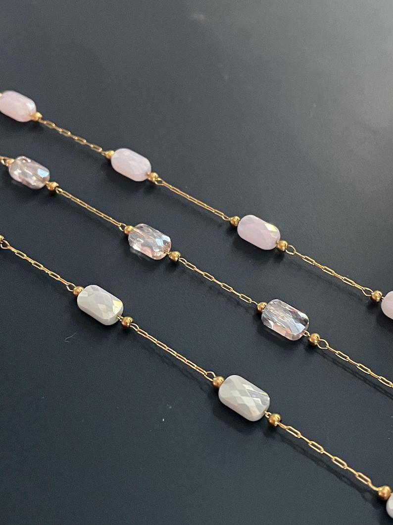 May include: Close-up of several delicate necklaces. Each necklace features a gold chain with alternating faceted, rectangular beads in shades of pink, clear, and white. Small gold beads accent the design, creating a refined and elegant look.