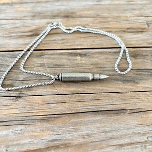 Puede incluir: Un collar de cadena de plata con un colgante de bala de plata. El collar est&aacute; sobre una superficie de madera.