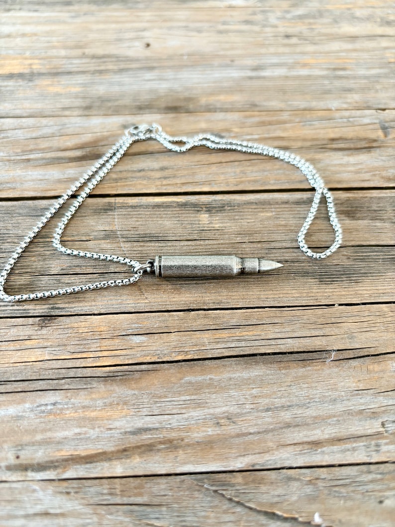 Puede incluir: Un collar de cadena de plata con un colgante de bala de plata. El collar est&aacute; sobre una superficie de madera.