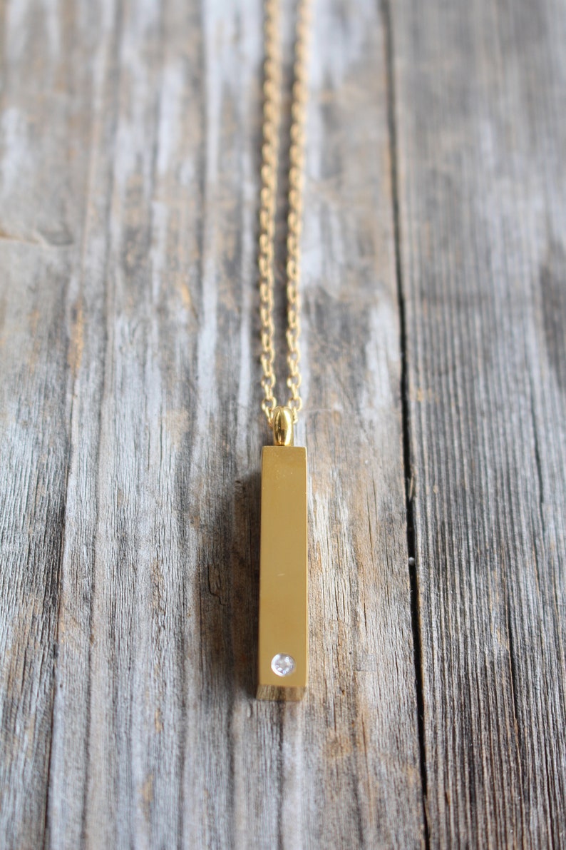 May include: A gold chain necklace with a gold bar pendant featuring a single clear rhinestone.