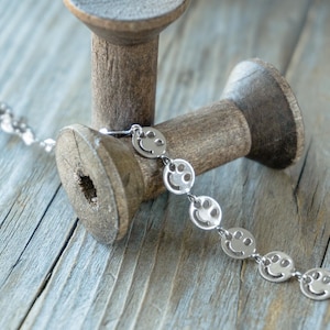 May include: A silver chain bracelet with a series of smiley face charms. The bracelet is lying on a wooden surface.