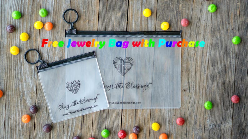 May include: Two clear plastic zippered bags with black zippers and black trim. The bags are on a wooden surface. The larger bag has the text "Shiny Little Blessings" printed on it. The smaller bag has the text "Free Jewelry Bag with Purchase" printed on it.