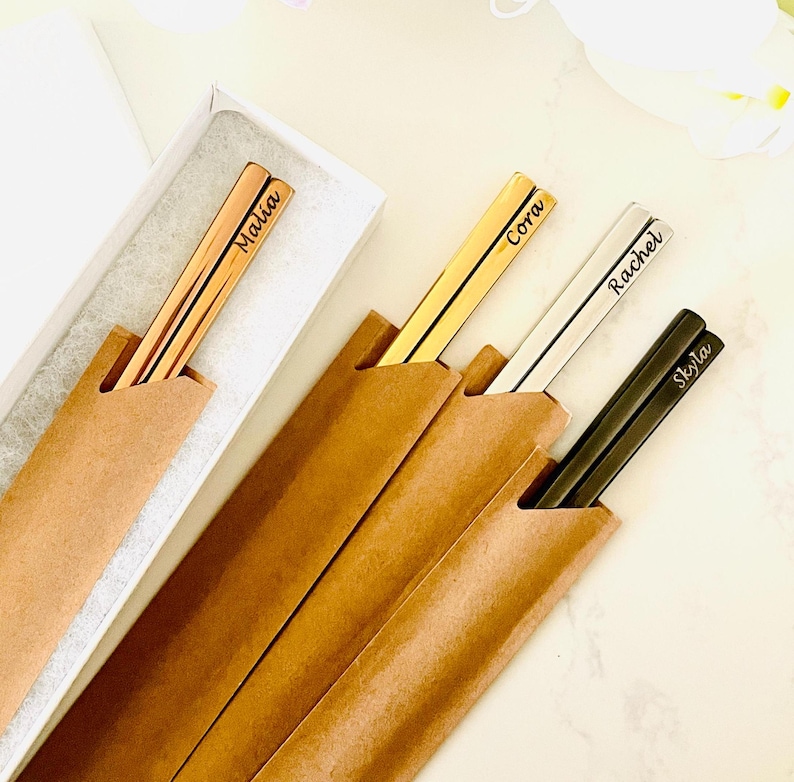 May include: Four sets of personalized chopsticks in brown paper packaging. The chopsticks are gold, silver, and black with the names Malia, Cora, Rachel, and Skyla engraved on them.