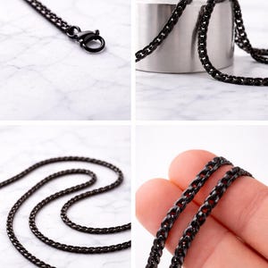 May include: Close-up images of a black chain necklace. The necklace, with small interlocking links, is shown with a lobster clasp. It's displayed on a white marble surface and a silver cylinder.