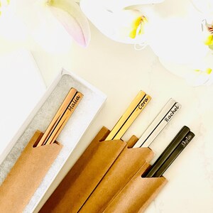 May include: Four sets of personalized chopsticks in brown paper sleeves. The chopsticks are gold, silver, and black with the names "Malia", "Cora", "Rachel", and "Skyla" engraved on them.