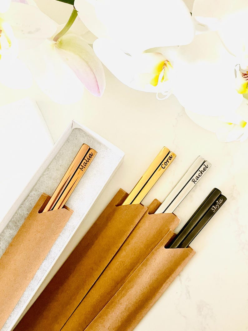 May include: Four sets of personalized chopsticks in brown paper sleeves. The chopsticks are gold, silver, and black with the names "Malia", "Cora", "Rachel", and "Skyla" engraved on them.