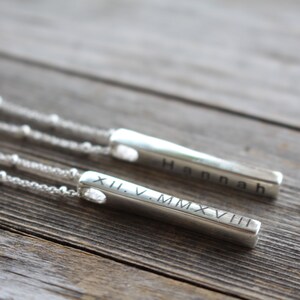 May include: Two silver bar necklaces with personalized engravings. One necklace has the name "Hannah" engraved on the bar, and the other has the Roman numeral date "XII.V.MMXVIII" engraved on the bar.