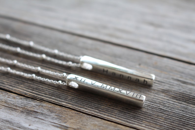 May include: Two silver bar necklaces with personalized engravings. One necklace has the name "Hannah" engraved on the bar, and the other has the Roman numeral date "XII.V.MMXVIII" engraved on the bar.