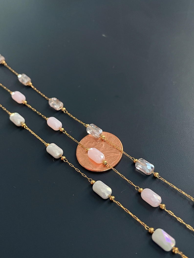 May include: Close-up of three delicate gold necklaces. Each necklace features a chain with alternating clear, pink, and white iridescent rectangular beads. A penny is included for scale, showing the necklaces' fine detail.