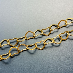 May include: Close-up of two gold chains featuring heart-shaped links. Each link has a textured, ridged surface. The chains are arranged diagonally against a dark grey background, providing a striking contrast.