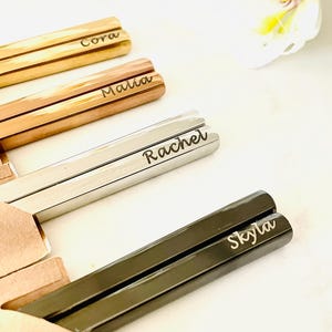 May include: Four pairs of chopsticks with personalized names: Cora, Malia, Rachel, and Skyla. The chopsticks are made of different materials, including gold, rose gold, silver, and black.