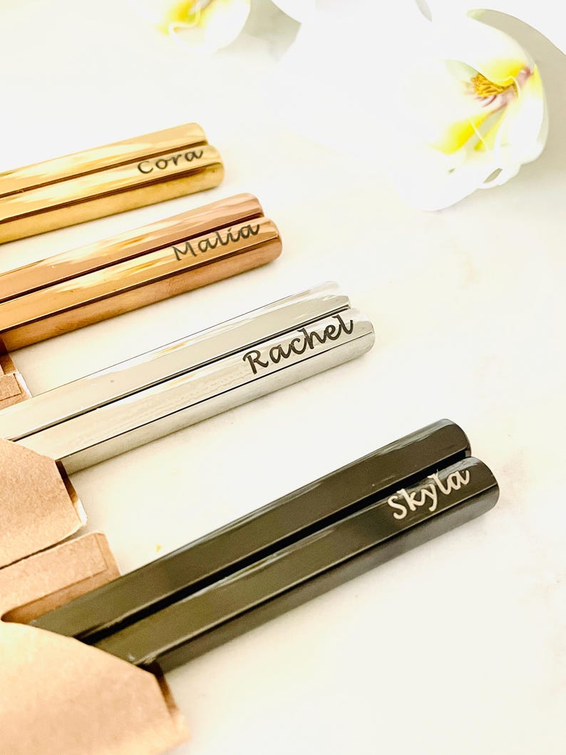 May include: Four pairs of chopsticks with personalized names: Cora, Malia, Rachel, and Skyla. The chopsticks are made of different materials, including gold, rose gold, silver, and black.