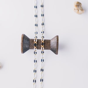 May include: Two white and black beaded necklaces with gold accents are wrapped around a wooden spool. The spool is on a white surface with several small white and brown stones.