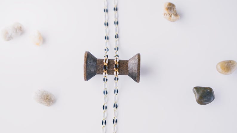 May include: Two white and black beaded necklaces with gold accents are wrapped around a wooden spool. The spool is on a white surface with several small white and brown stones.