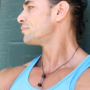 May include: A man wearing a light blue tank top with a black chain necklace and a clear glass pendant. The pendant has a black metal top and bottom.