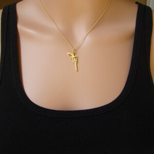 Pode incluir: Colar com pingente dourado com um berloque de rev&oacute;lver detalhado. O berloque pende de uma corrente delicada. O colar &eacute; usado sobre uma regata preta.