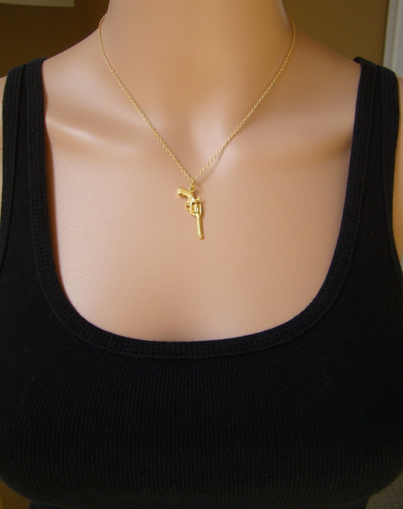 Pode incluir: Colar com pingente dourado com um berloque de rev&oacute;lver detalhado. O berloque pende de uma corrente delicada. O colar &eacute; usado sobre uma regata preta.