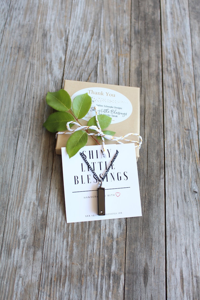 May include: A black necklace with a small rectangular pendant that says "Shiny Little Blessings" on a white card. The card also says "Handmade with love" and includes a website address.