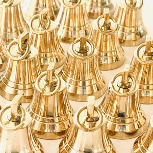 May include: A group of 15 small, shiny, gold-colored bells with a decorative handle on top. The bells are arranged in a semi-circle on a white surface.